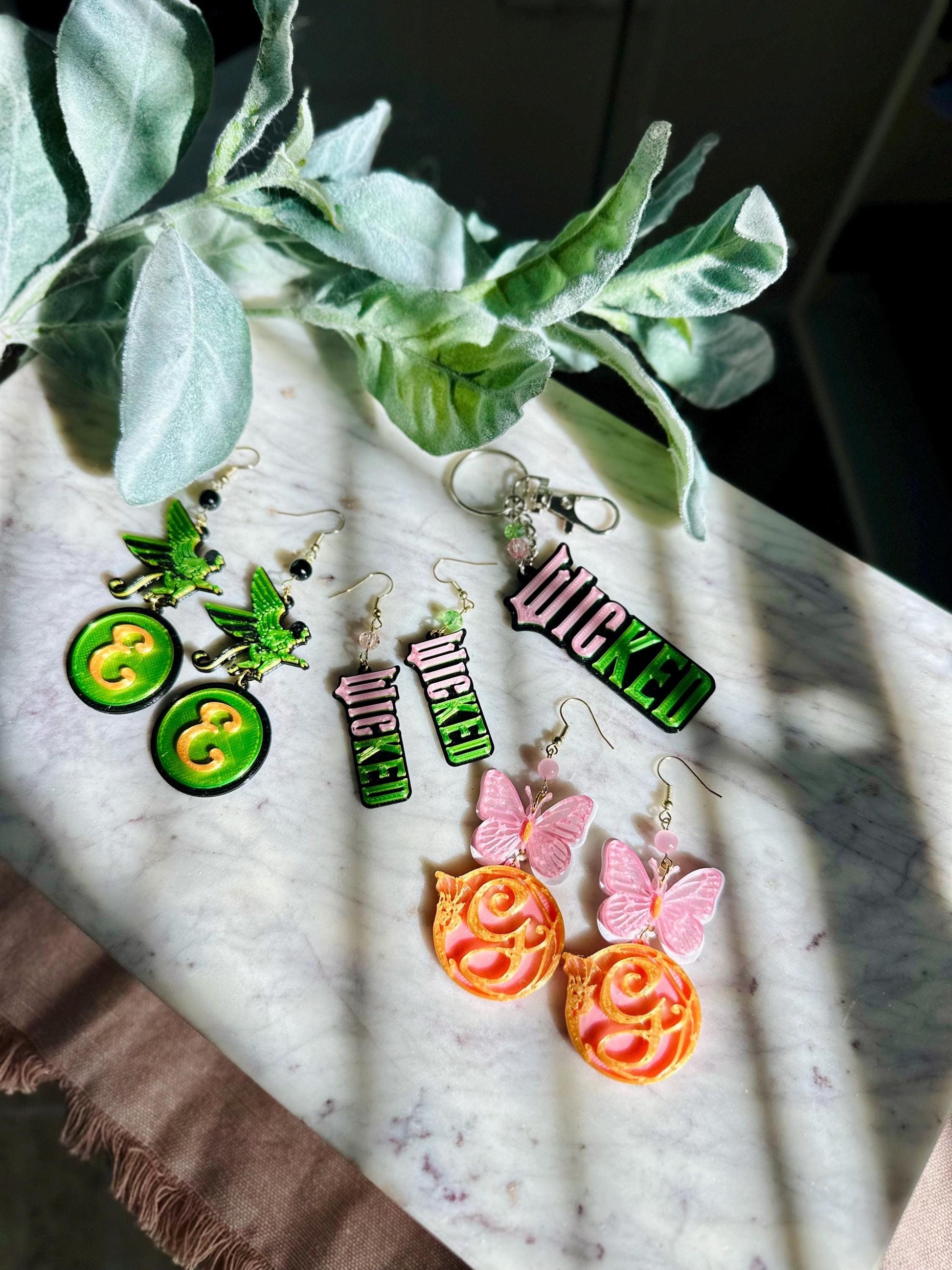 Wicked earrings, Green monkey tiered with Elphaba logo. Butterfly paired with gold pink Glinda Shiz logo