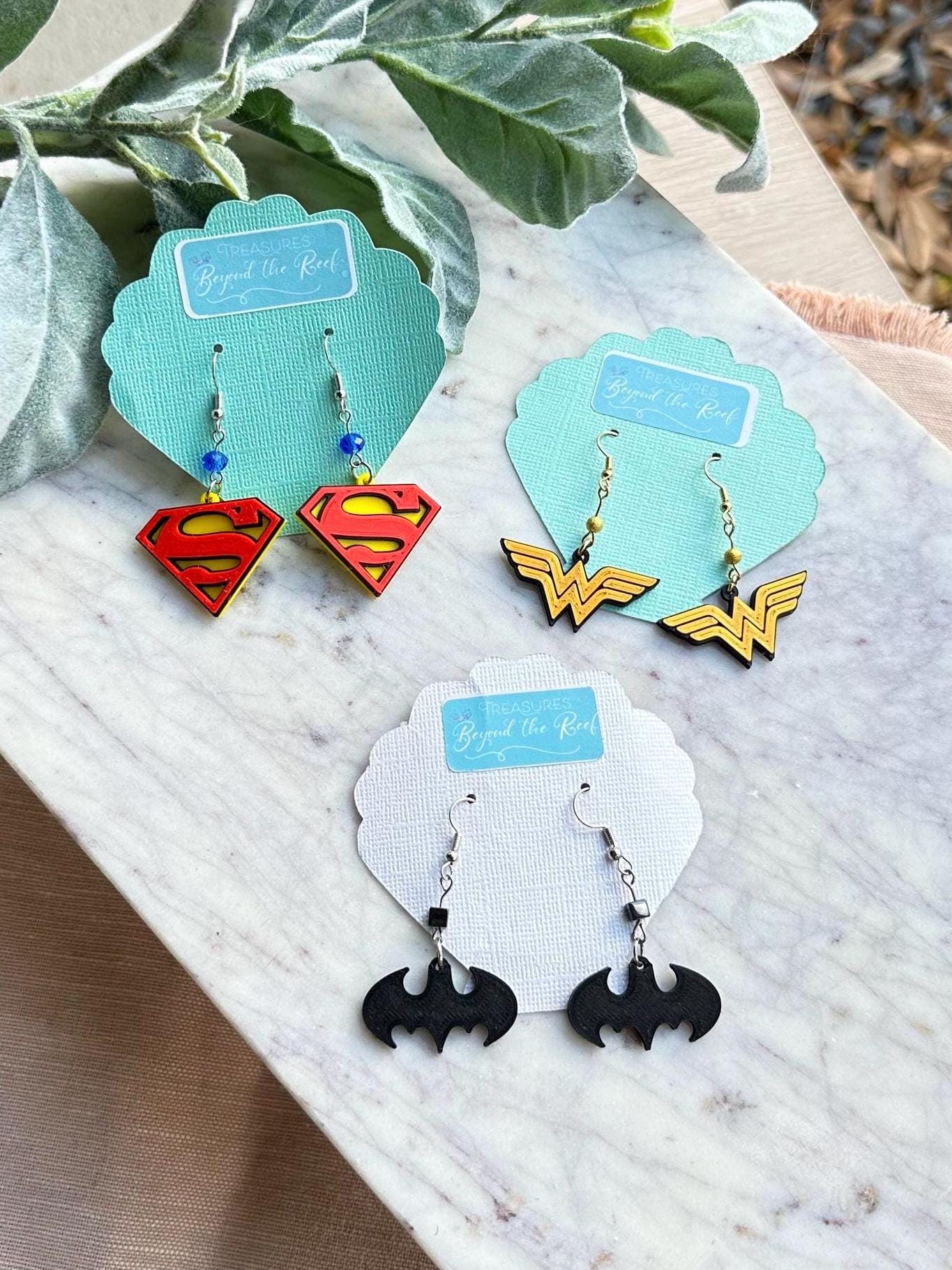 May include: Three pairs of superhero themed earrings on white and blue display cards. The earrings are silver with black, red, yellow, and blue accents. The cards have the words &#39;Treasures Beyond the Reef&#39; printed on them.