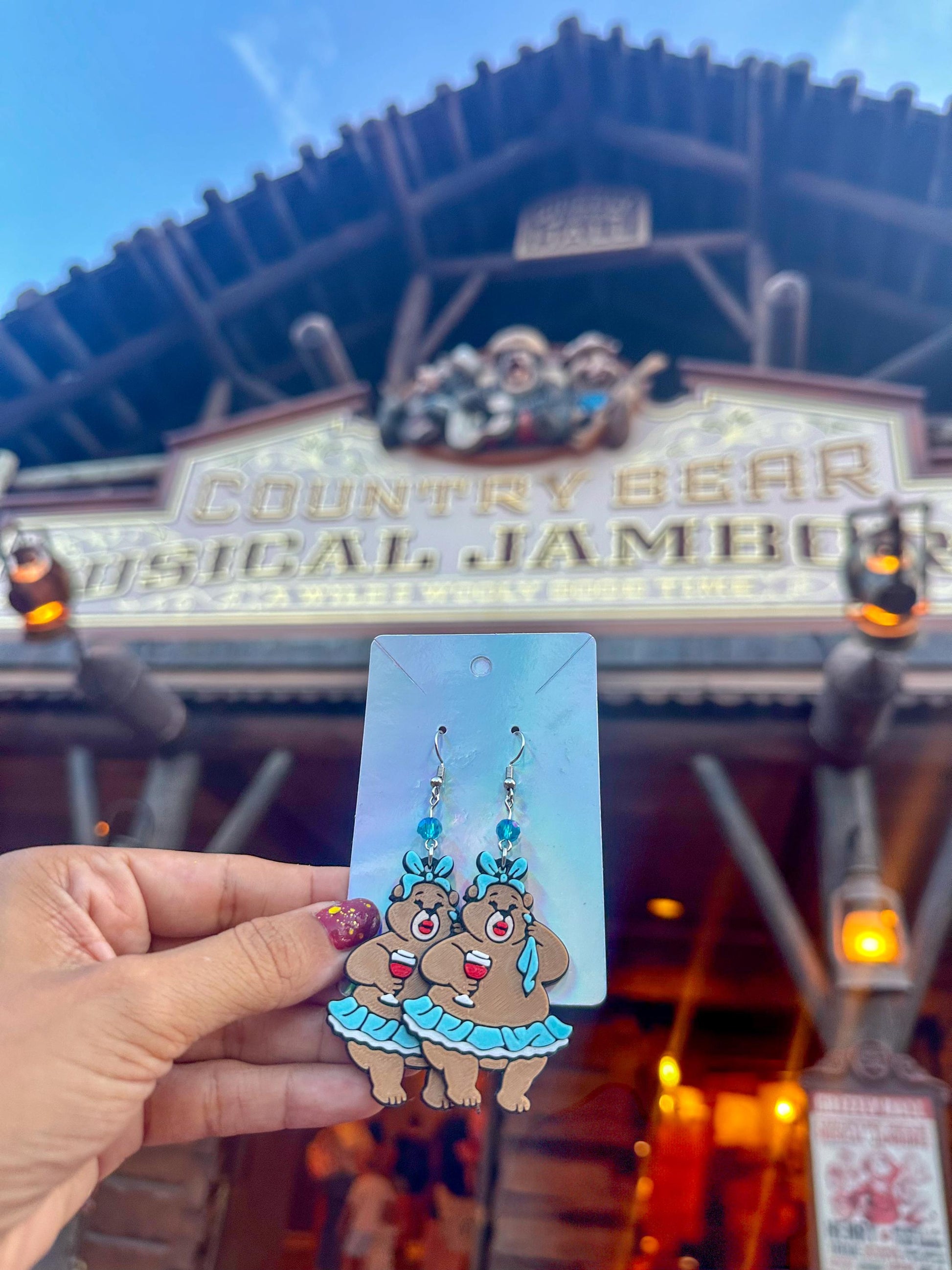 May include: A pair of dangle earrings featuring cartoon bears holding wine glasses. The bears are brown with blue skirts and bows. The earrings have blue beads and silver hooks. The background shows the Country Bear Jamboree sign.