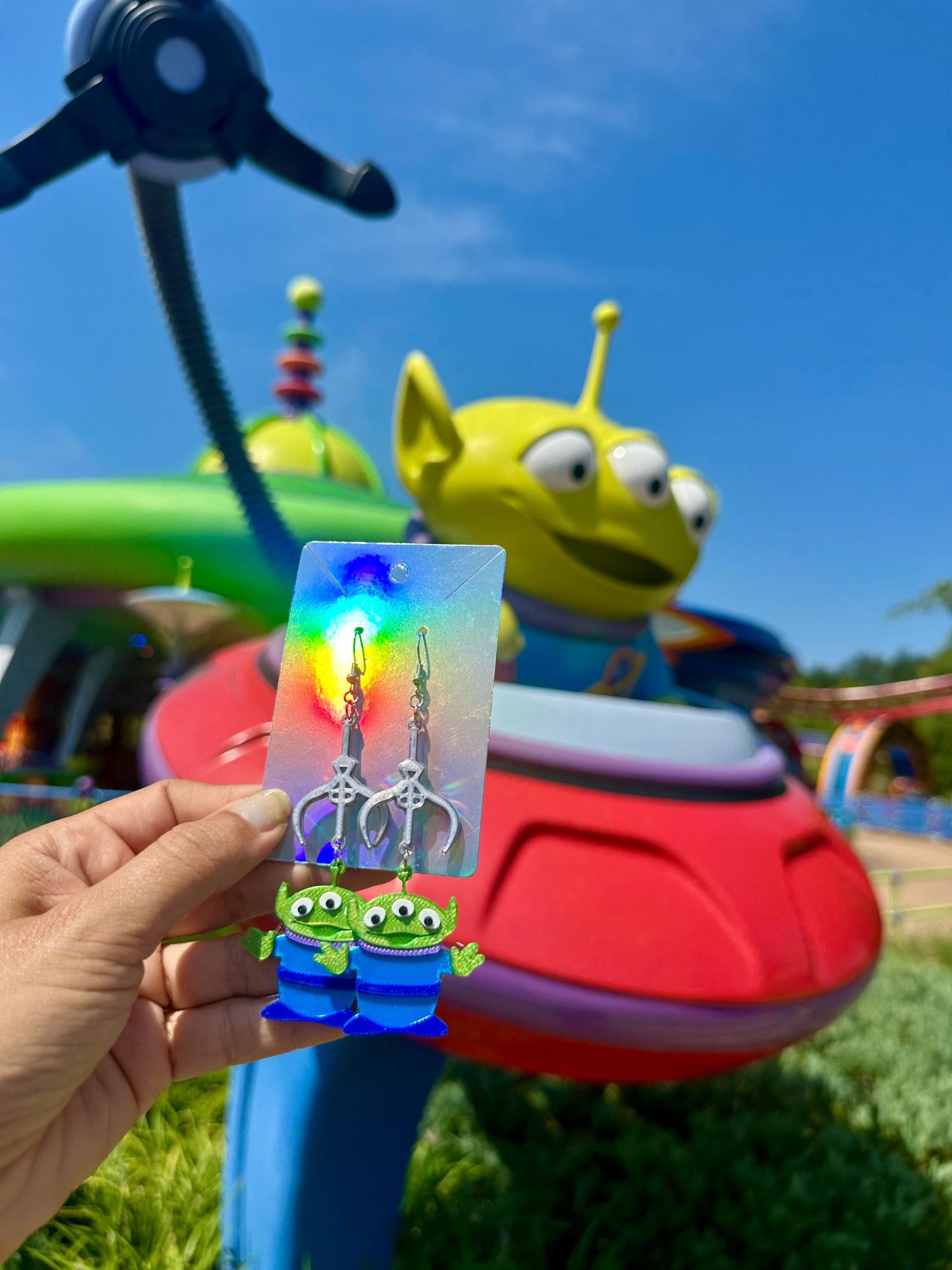 May include: Alien-themed earrings featuring the claw from Toy Story. The earrings have silver claw charms and green alien charms with three eyes and blue striped outfits. The earrings are displayed in front of a Toy Story alien ride with a blue sky background.
